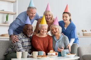 Group portrait of happy multiracial senior people and young woman nurse celebrating attractive elderly lady birthday at nursing home, sitting on couch, looking at birthday cake and smiling